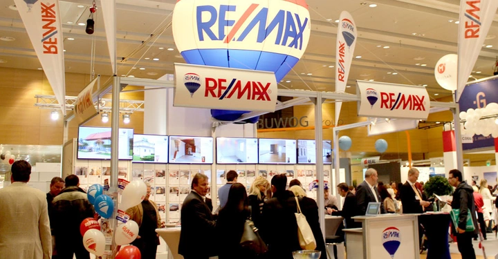 Galerie-Bild 13: Luftballon mit RE/MAX-Logo schwebt über einem belebten Messestand mit mehreren Bildschirmen; Menschen interagieren mit dem Stand, auf dem weiteres RE/MAX-Material ausgestellt ist.