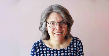 Eine Frau mit grauem Haar und Brille lächelt leicht vor einem hellrosa Hintergrund Sie trägt ein blaues Oberteil mit Blumenmuster