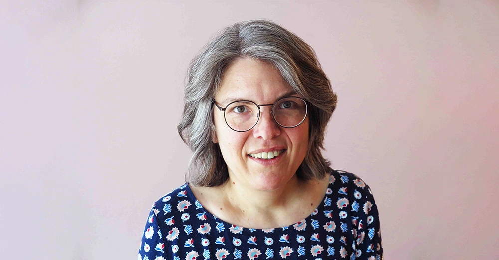 Eine Frau mit grauem Haar und Brille lächelt leicht vor einem hellrosa Hintergrund Sie trägt ein blaues Oberteil mit Blumenmuster