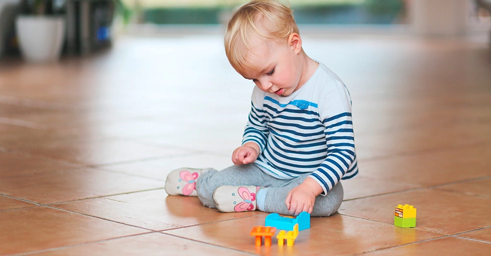 Kleinkind spielt auf einem Fliesenboden mit bunten Spielzeugklötzen in einem hellen Raum mit Topfpflanzen im Hintergrund.