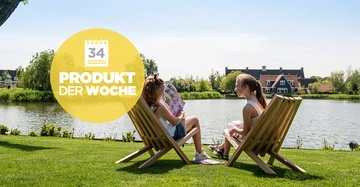 Zwei Personen sitzen auf Holzstühlen und lesen Zeitschriften am Ufer eines Flusses mit Häusern im Hintergrund. Text: Kalender mit Nummer 34 PRODUKT DER WOCHE