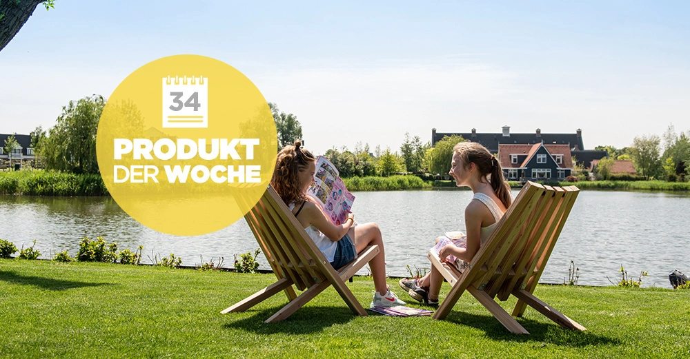 Zwei Personen sitzen auf Holzstühlen und lesen Zeitschriften am Ufer eines Flusses mit Häusern im Hintergrund. Text: Kalender mit Nummer 34 PRODUKT DER WOCHE