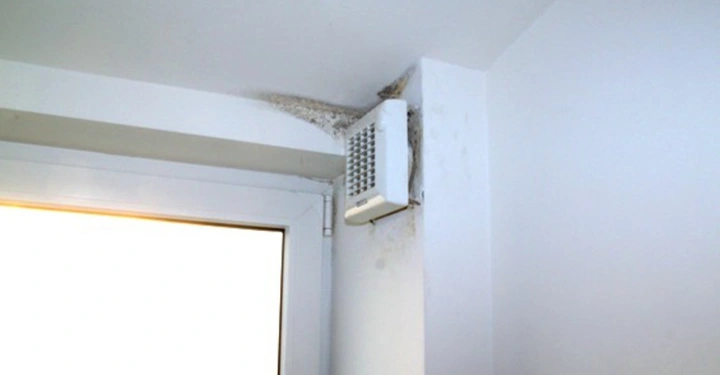 Galerie-Bild 16: Ein weißer Lüftungsschacht ist an einer Zimmerdecke befestigt Schimmelbildung ist um den Schacht herum sichtbar Die Wand und Decke sind weiß gestrichen und es gibt ein Fenster daneben