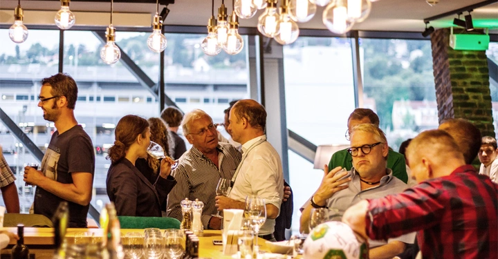 Galerie-Bild 6: Personen unterhalten sich an einer Bar in einem modernen Raum mit großen Fenstern und Glühbirnen über dem Tresen. Ein Mann hält ein Smartphone, ein anderer Mann sitzt mit einem Fußball.