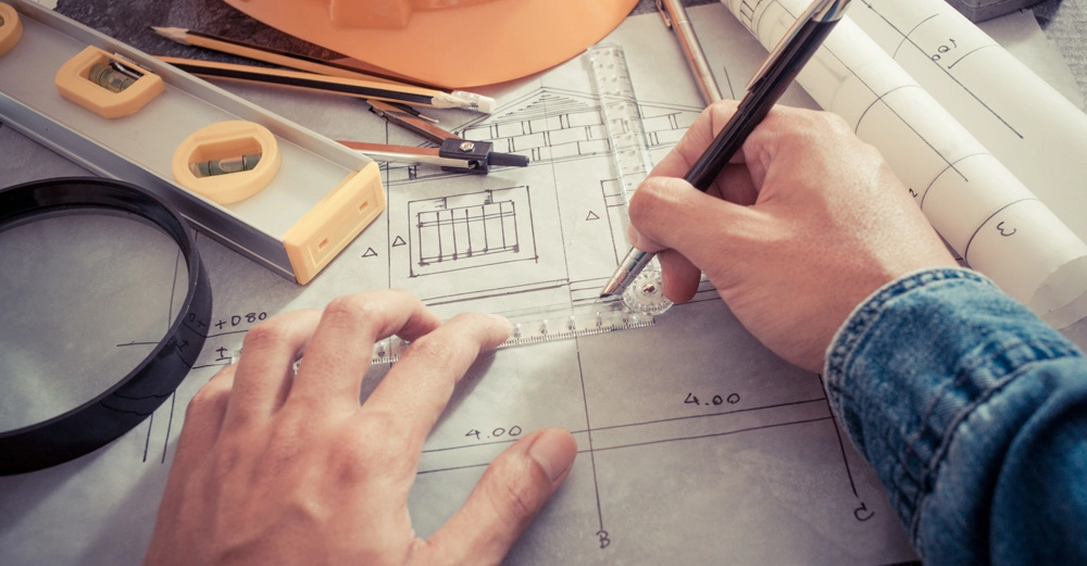 Vogelperspektive eines Schreibtischs, auf dem ein Mann einen Bauplan bearbeitet. Helm, Wasserwaage, Stifte und Maßband sind auf dem Tisch verstreut.