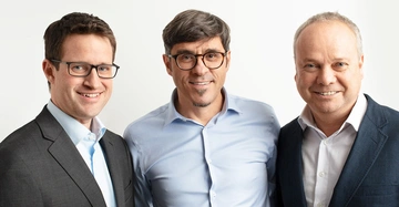 Drei Männer stehen nebeneinander und lächeln in die Kamera alle tragen Hemden und Anzüge. Der Hintergrund ist vor einem weißen Hintergrund.