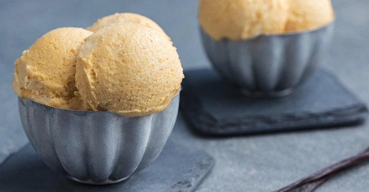 Drei Kugeln Vanilleeis befinden sich in einer silbernen Schüssel auf einem dunklen Untersetzer. Im Hintergrund steht eine weitere Schüssel mit drei Kugeln Vanilleeis.