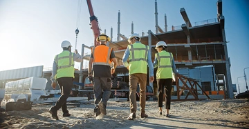 Vier Bauarbeiter gehen auf einer Baustelle mit einem im Bau befindlichen Gebäude im Hintergrund. Sie tragen Schutzhelme und reflektierende Westen. Ein Kran arbeitet im Hintergrund.