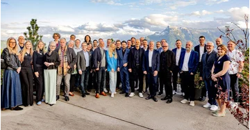 große Gruppe von Männern und Frauen im Businesslook steht auf sonniger Terrasse vor Bergkulisse