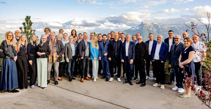 große Gruppe von Männern und Frauen im Businesslook steht auf sonniger Terrasse vor Bergkulisse