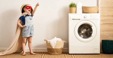 Ein Kind trägt eine rote Maske und einen Umhang hebt triumphierend die Faust im Zimmer in dem eine Waschmaschine und Wäschekörbe stehen.