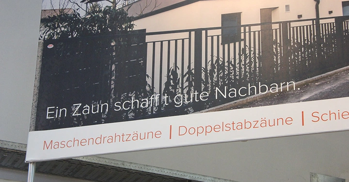 Galerie-Bild 21: Ein eiserner Zaun umgibt ein modernes Haus beleuchtet in der Dämmerung mit dem Text Ein Zaun schafft gute Nachbarn. Unten steht Maschendrahtzäune Doppelstabzäune Schiebetore in oranger Schrift.