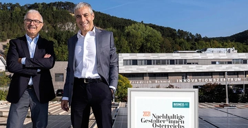 Zwei Männer posieren lächelnd in Anzügen. Im Hintergrund befindet sich das Friedrich Schmid Innovationszentrum vor einer bewaldeten Landschaft. Text: 2021 Nachhaltige Gestalter*innen Österreichs BUSINESS ART