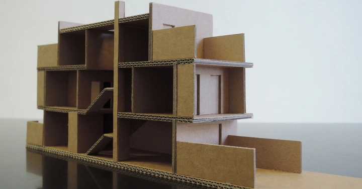 Kartonskulptur zeigt ein mehrstöckiges Gebäude mit verschiedenen Räumen und Treppen auf einem glatten Tisch in einem minimalistischen Raum