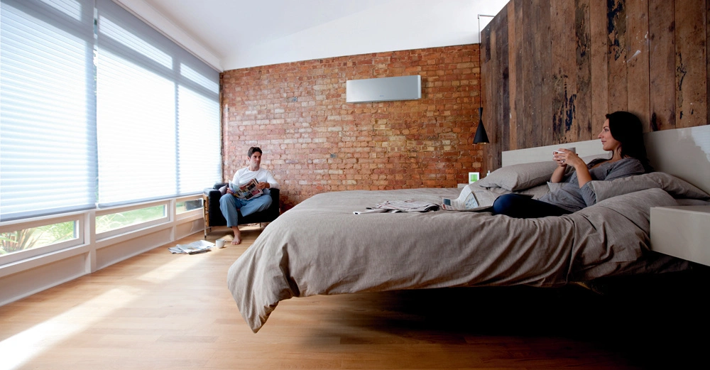 Zwei Personen entspannen in einem modernen Schlafzimmer. Eine Person sitzt in einem Sessel und liest ein Buch, während die andere Person auf einem Hochbett sitzt und eine Tasse hält.