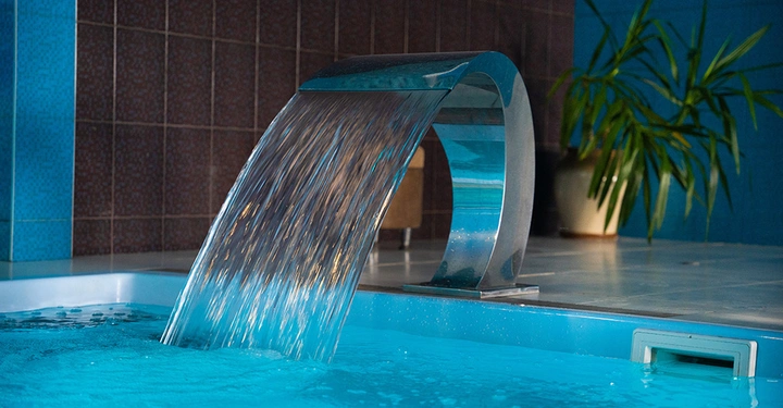 breite Edelstahl-Wasserdusche am Rand eines Indoorpools, die gerade läuft. Poolraum ist abendlich in blauen Farbtönen, sieht sehr elegant und gemütlich aus