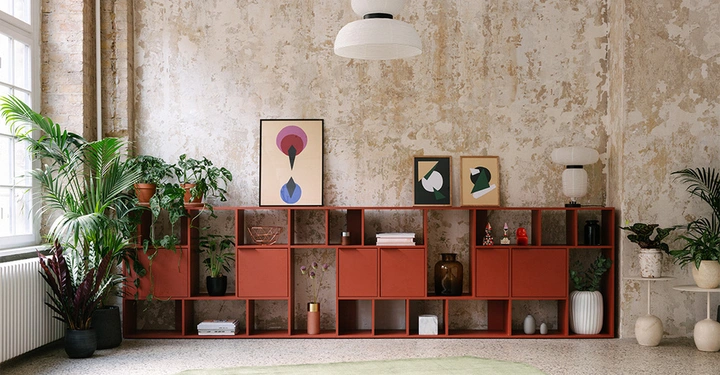 Ein großes, rotes Regal steht gegen eine abgenutzte Wand es enthält verschiedene Pflanzen und dekorative Gegenstände wie Vasen und Bilderrahmen der Raum ist mit Pflanzen und einem Teppich dekoriert.