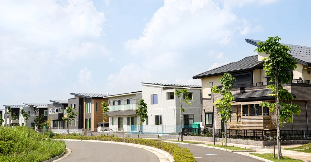 Moderne zweistöckige Häuser stehen ruhig in einer neugebauten Wohngegend neben frischen Bäumen und einem gebogenen Gehweg unter einem wolkigen Himmel.