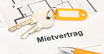 Mietvertrag liegt auf Tisch mit Kuli zum Unterschreiben, Wohnungsschlüssel mit Anhänger liegt darauf, im Hintergrund ein Grundrissplan