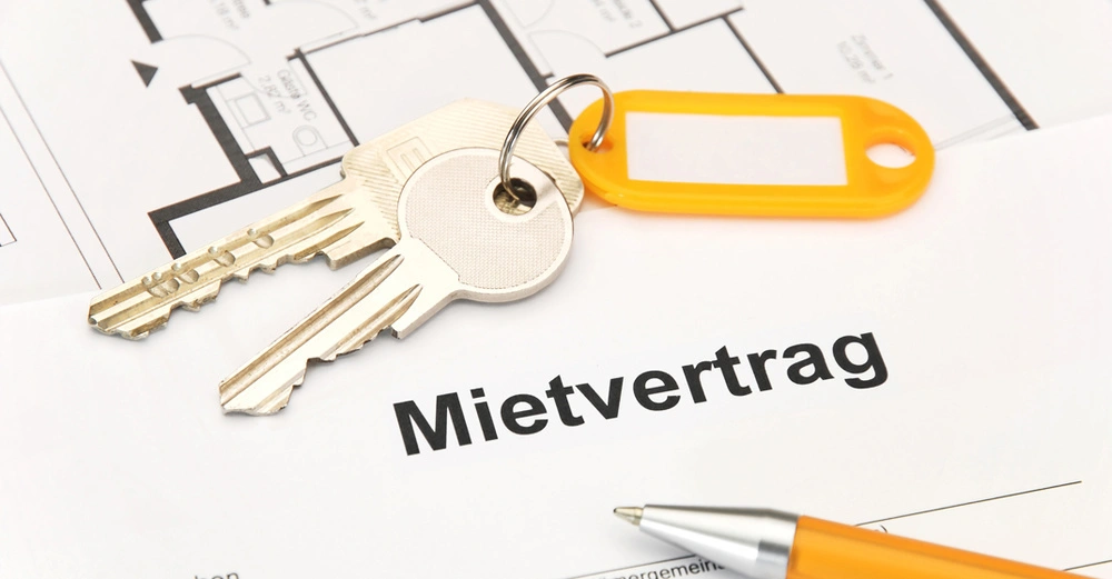 Mietvertrag liegt auf Tisch mit Kuli zum Unterschreiben, Wohnungsschlüssel mit Anhänger liegt darauf, im Hintergrund ein Grundrissplan