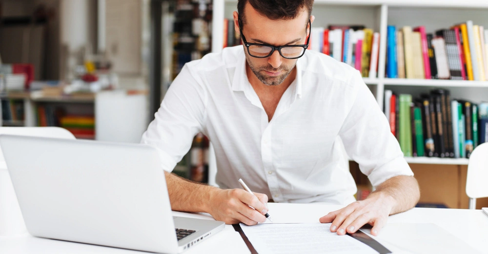 Ein Mann sitzt an einem Schreibtisch und schreibt auf ein Dokument; neben ihm steht ein offener Laptop; im Hintergrund befinden sich Bücherregale in einem Bürozimmer.