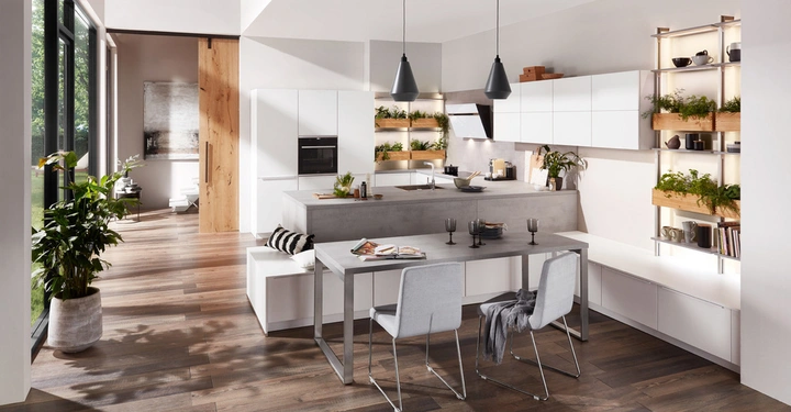 Moderne Küche mit weißen Schränken und grauer Arbeitsplatte Töpfe und Pflanzen dekorieren die Regale Große Fenster lassen viel Licht herein Parkettboden und ein Holztisch mit zwei grauen Stühlen stehen im Vordergrund