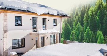 Ein zweistöckiges Haus mit Flachdach ist von Schnee bedeckt. Im Hintergrund befinden sich üppige grüne Nadelbäume.