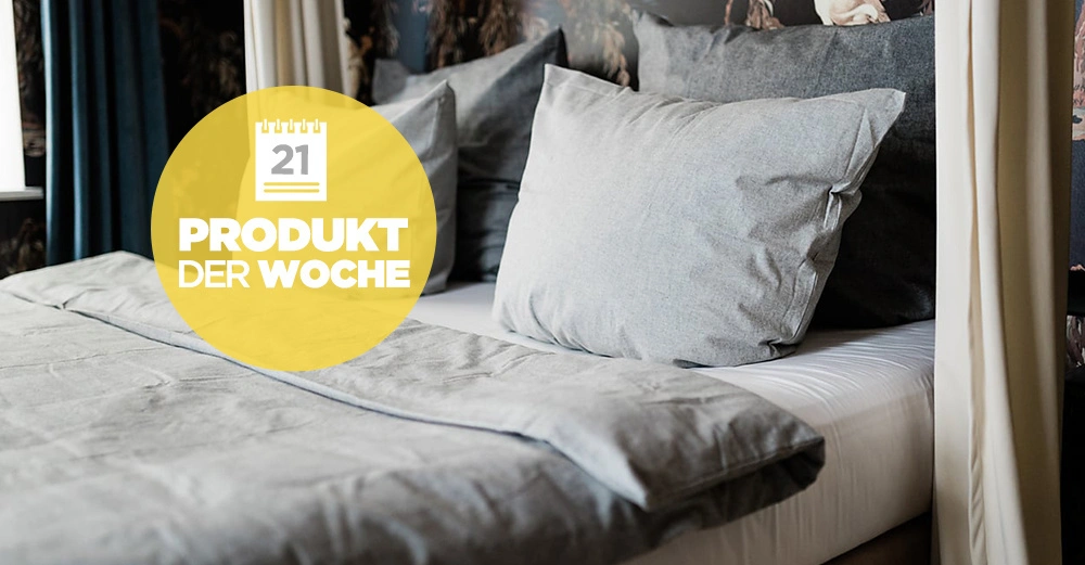 Graues Bettlaken und Kissen auf einem Bett umgeben von Vorhängen und dunkel gemusterten Wänden gelber Kreis mit Text 21 PRODUKT DER WOCHE.