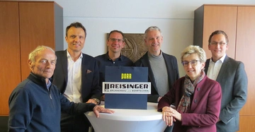 Sechs Personen stehen um einen runden Tisch in einem Büro und lächeln. Auf dem Tisch befindet sich ein Schild mit der Aufschrift DORR REISINGER BRUNNENBAU UND BOHRTECHNIK.