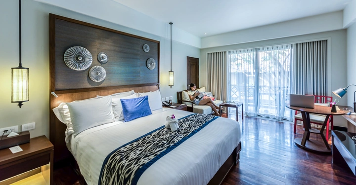 Ein gemütliches Schlafzimmer mit einem großen Bett dekoriert mit blauen Kissen und einem kunstvollen Holzbrett im Hintergrund Eine Person liest auf einem Sofa nahe einem großen Fenster mit Vorhängen