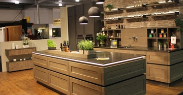 Großer Kücheninsel-Tresen ausgestattet mit Kräutertöpfen und Flaschen stehend auf einem Holzboden vor einer braunen Steinwand mit Wandregalen unter dunklen Lampen in einer modernen Küchenumgebung