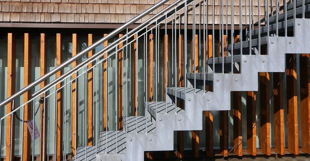 Außentreppe aus Beton führt an Haus mit Holzfassade hoch. Hochwertiges Geländer mit Handlauf aus silbernem Metall schließt die Treppenwange ab. 
