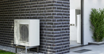 Eine weiße Klimaanlage steht an einer grauen Ziegelwand, daneben befindet sich eine offene Tür zu einem modernen Raum und links ist grüner Rasen zu sehen.