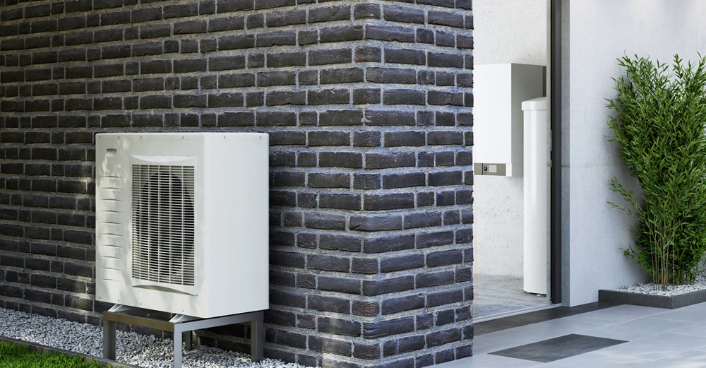 Eine weiße Klimaanlage steht an einer grauen Ziegelwand, daneben befindet sich eine offene Tür zu einem modernen Raum und links ist grüner Rasen zu sehen.