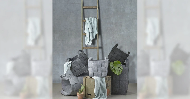Galerie-Bild 4: Gewebte graue Körbe stehen gestapelt auf einem Betonboden, einige enthalten gezackte Decken und eine Monstera-Pflanze. Daneben befindet sich eine hölzerne Leiter mit einer hängenden Decke vor einer grauen Wand.