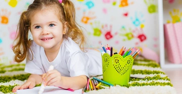 Ein Mädchen malt auf dem Boden liegend mit einem Stift aus einem grünen Behälter in einem bunt dekorierten Raum voller Handabdrücke an der Wand.