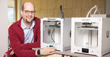 freundlich lächelnder Mann mit Halbglatze und rahmenloser, runder Brille in roter Strickjacke sitzt vor zwei weißen Geräten namens Ulitmaker.