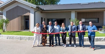 Eine Gruppe von neun Menschen schneidet ein rotes Band vor einem modernen Gebäude durch. Alle lächeln und stehen auf einem Gehweg bei sonnigem Wetter mit Rasenflächen und Bäumen im Hintergrund.