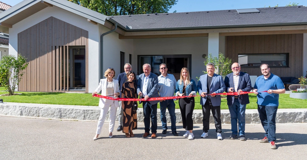 Eine Gruppe von neun Menschen schneidet ein rotes Band vor einem modernen Gebäude durch. Alle lächeln und stehen auf einem Gehweg bei sonnigem Wetter mit Rasenflächen und Bäumen im Hintergrund.
