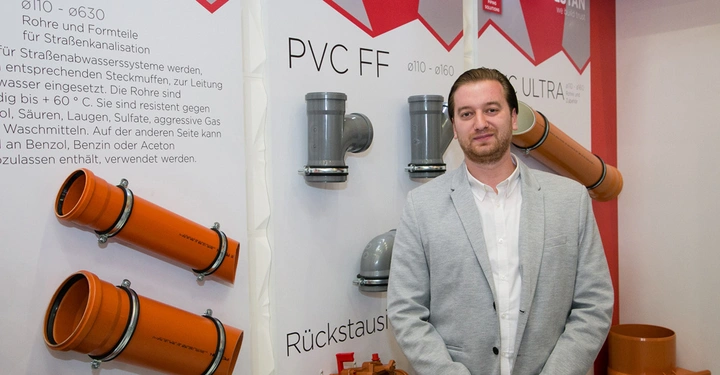 Galerie-Bild 8: Ein Mann in einem grauen Anzugjackett steht vor einer Ausstellungswand mit orangefarbenen und grauen PVC-Rohren. An der Wand sind verschiedene Beschreibungen zu den Rohren zu sehen. Text: ø110-ø630 Rohre und Formteile für Straßenkanalisation...