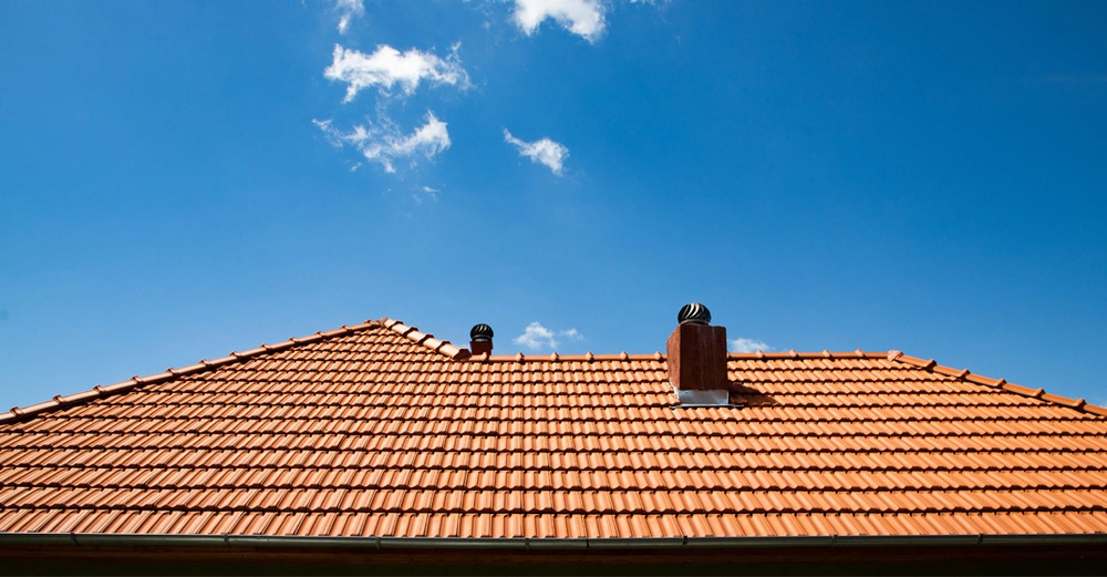 rotes Ziegeldach mit 2 Kaminen vor strahlend blauem Himmel mit kleinen Wölkchen