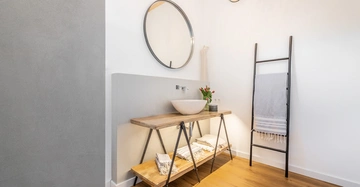 Waschbecken auf einem Holztisch mit einem runden Spiegel darüber befindet sich in einem Badezimmer. Rechts daneben eine Leiter mit einem Handtuch. Logo "LAMURISTA" unten rechts.