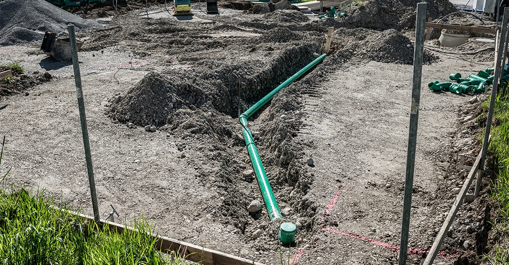 Grünes Rohr liegt in Erdgraben auf Baustelle umgeben von Erdhaufen und Baumaterialien bei sonnigem Wetter.