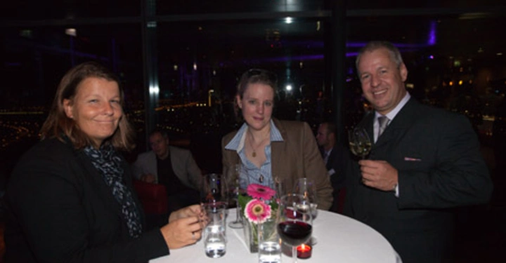 Galerie-Bild 79: Drei Personen stehen an einem runden Tisch und halten Getränke in der Hand dunkel beleuchteten Raum im Hintergrund weitere sitzende Menschen. Blumen und Gläser stehen auf dem Tisch.