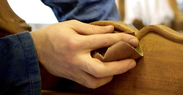 Eine Hand schleift ein Holzstück in einer Werkstatt ab die Person trägt blaue Arbeitskleidung und die Umgebung ist unscharf