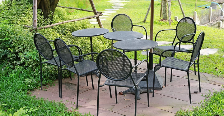 Schwarze Metallstühle und runde Tische stehen auf einer gepflasterten Terrasse umgeben von üppigem grünem Garten und einem Steinweg im Hintergrund.