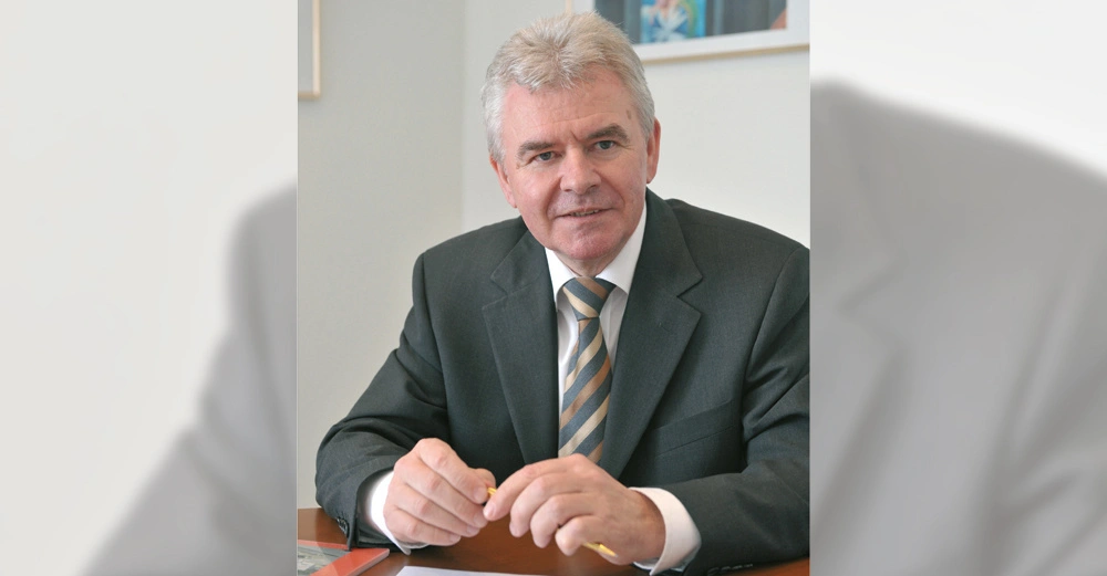 Ein Mann im grauen Anzug sitzt an einem Schreibtisch mit Papier und hält einen Stift in der Hand, während er in die Kamera schaut, in einem Büro mit Wandbildern.