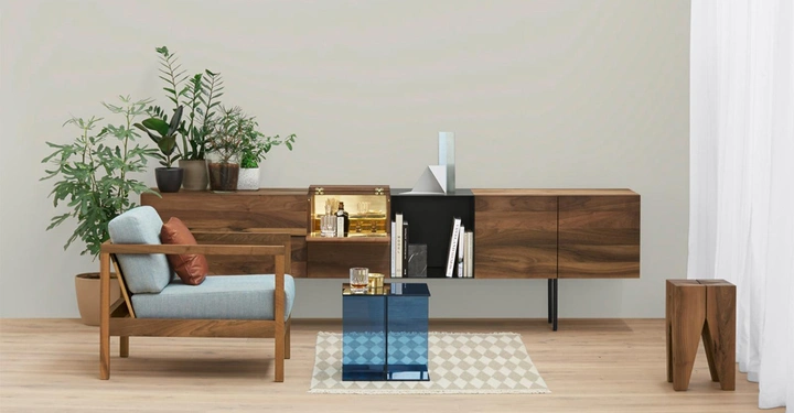 Ein blauer gläserner Couchtisch steht vor einem Holzstuhl mit blauen Kissen und orangefarbenem Kissen im Wohnzimmer mit Holzschränken an der Wand und vielen grünen Pflanzen.