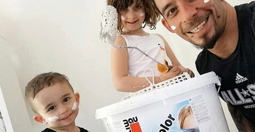 Mann und zwei Kinder malen zusammen eine Wand mit weißer Farbe. Sie lächeln und haben Farbflecken im Gesicht. Der Mann hält einen Farbeimer.