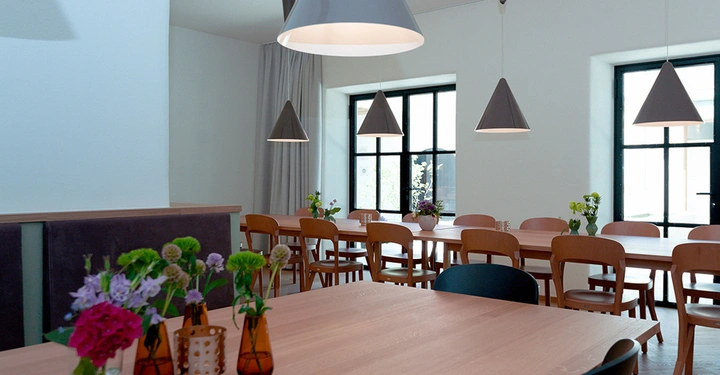 Galerie-Bild 3: Holztische mit Blumenvasen stehen unter hängenden Lampen in einem hellen Raum mit Holzwand und großen Fenstern. Im Hintergrund befinden sich weitere Tische und Stühle.
